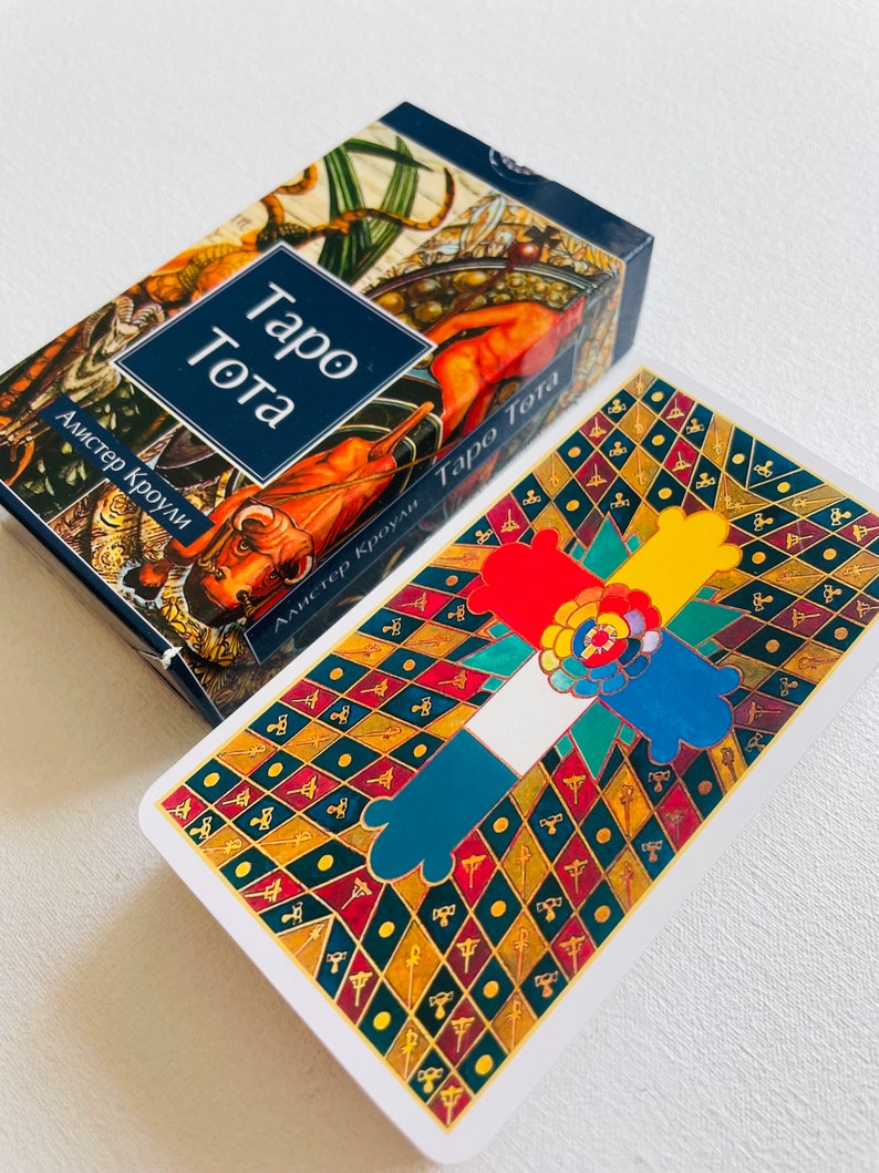 Pu&ograve; includere: Una scatola di mazzo di tarocchi con uno sfondo blu e un'illustrazione colorata di un toro e un uomo a cavallo. Il testo "Taro Tota" &egrave; stampato in bianco su uno stendardo blu. Il testo "Alister Crowley" &egrave; stampato in bianco su uno stendardo blu. La scatola &egrave; aperta ed &egrave; visibile una carta dei tarocchi. La carta ha uno sfondo bianco con un design colorato di una croce a quattro braccia, ogni braccio di un colore diverso. La croce &egrave; circondata da uno schema di diamanti neri e dorati.