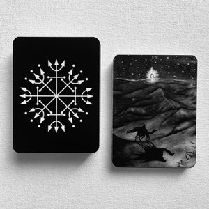 Może przedstawiać: Dwie czarne karty tarota z białymi wzorami. Karta po lewej stronie ma biały symbol ze strzałkami skierowanymi na zewnątrz. Karta po prawej stronie przedstawia czarno-białą ilustrację wielbłąda idącego przez pustynny krajobraz z rozgwieżdżonym niebem nad nim.