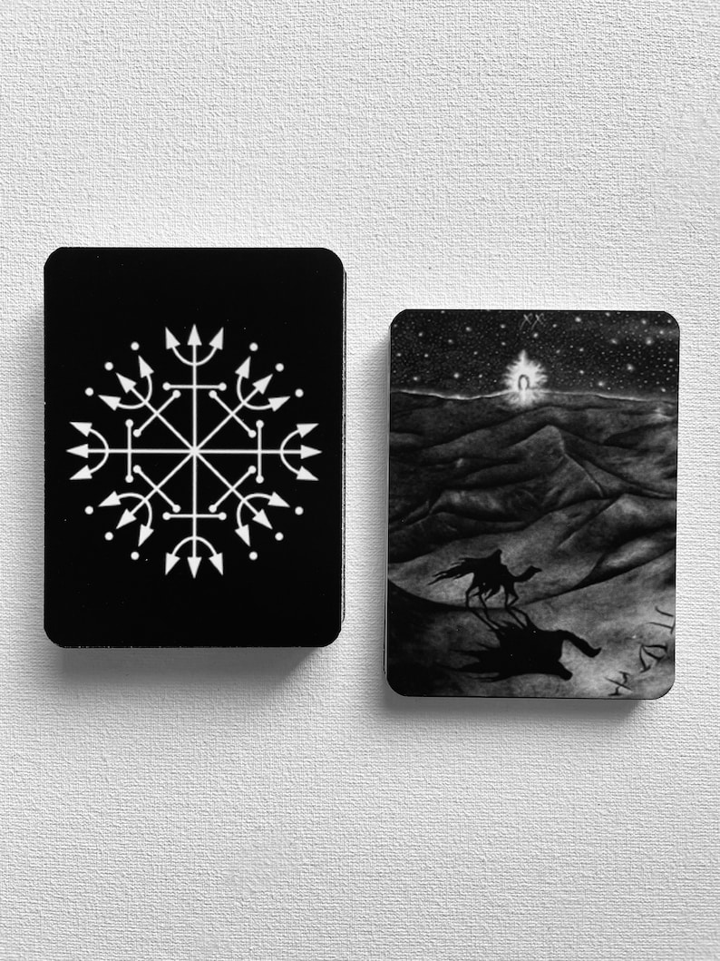 Może przedstawiać: Dwie czarne karty tarota z białymi wzorami. Karta po lewej stronie ma biały symbol ze strzałkami skierowanymi na zewnątrz. Karta po prawej stronie przedstawia czarno-białą ilustrację wielbłąda idącego przez pustynny krajobraz z rozgwieżdżonym niebem nad nim.