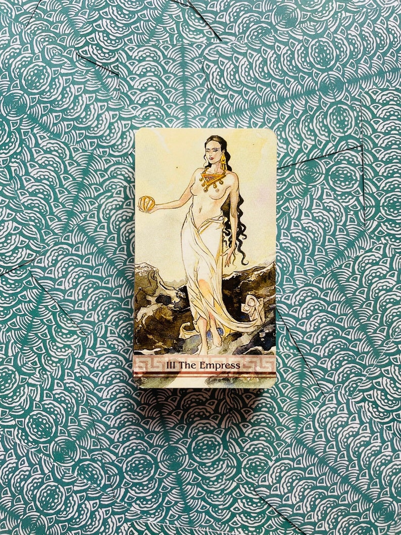 May include: The Empress tarot card features a woman with long dark hair, wearing a white robe and gold jewelry. She holds a golden orb in her left hand and stands on a rocky outcrop. The card is titled "III The Empress" in a red banner.