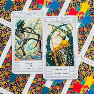 Pu&ograve; includere: Due carte dei tarocchi, Morte e Regina di Denari, con sfondo bianco e bordo argentato. La carta della Morte presenta una figura scheletrica in un paesaggio blu e bianco. La carta della Regina di Denari presenta una donna con una corona e uno scettro, seduta su un trono con un ariete ai suoi piedi.