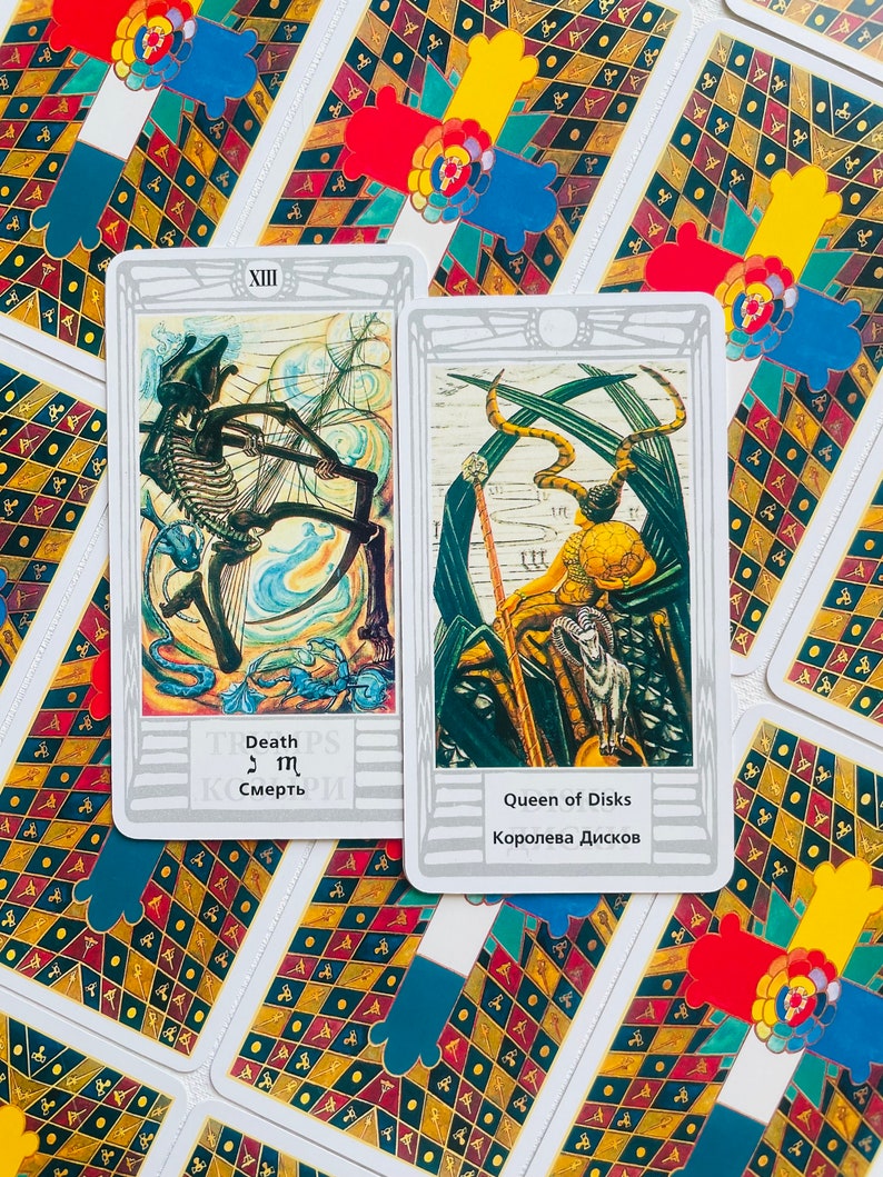 Pu&ograve; includere: Due carte dei tarocchi, Morte e Regina di Denari, con sfondo bianco e bordo argentato. La carta della Morte presenta una figura scheletrica in un paesaggio blu e bianco. La carta della Regina di Denari presenta una donna con una corona e uno scettro, seduta su un trono con un ariete ai suoi piedi.