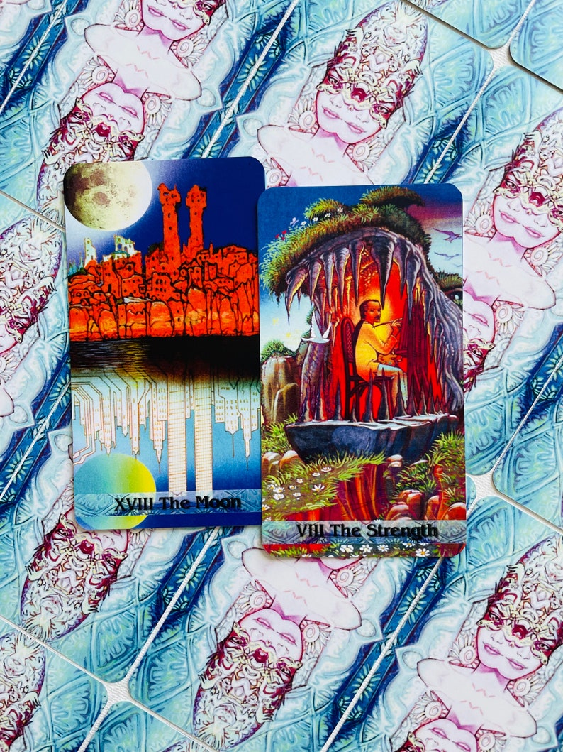 K&ouml;nnte beinhalten: Zwei Tarotkarten, Der Mond und Die St&auml;rke, mit bunten Illustrationen. Die Mondkarte zeigt eine Skyline mit einem Vollmond am Himmel. Die St&auml;rkekarten zeigt eine Person, die in einer H&ouml;hle mit einem L&ouml;wen sitzt.