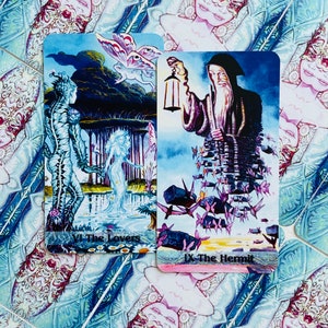 K&ouml;nnte beinhalten: Zwei Tarotkarten, Die Liebenden und Der Einsiedler, mit farbenfrohen Illustrationen. Die Karte Die Liebenden zeigt eine blaue und gr&uuml;ne Kreatur mit einem Schmetterling, w&auml;hrend die Karte Der Einsiedler eine Figur in einem violetten Gewand zeigt, die eine Laterne h&auml;lt.