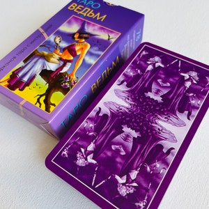 Peut inclure: Une carte de tarot violette avec le texte "Taro Vedm" sur le devant. La carte pr&eacute;sente un fond violet fonc&eacute; avec trois personnages en longues robes et un motif floral.