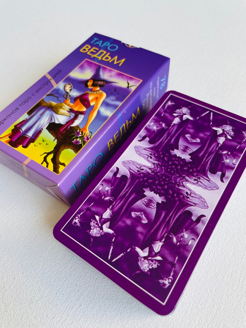Peut inclure: Une carte de tarot violette avec le texte "Taro Vedm" sur le devant. La carte pr&eacute;sente un fond violet fonc&eacute; avec trois personnages en longues robes et un motif floral.