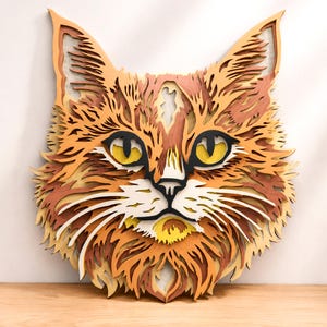 May include: A multi-layered wooden wall art piece shaped like a cat head. The design features intricate cutouts in shades of orange, tan, brown, and white, with bright yellow eyes and black accents, creating a dimensional effect of a cat face.