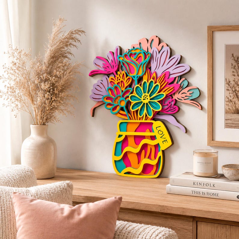 May include: A multi-layered wooden wall art piece depicting a bouquet of stylized flowers in a vase. The flowers are colored in shades of pink, purple, orange, and teal. The vase is yellow with wavy lines and a tag that reads the word LOVE.