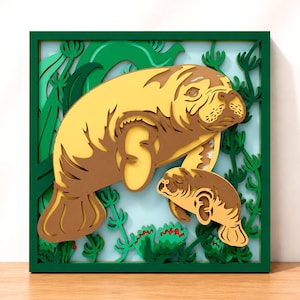 May include: A square, multi-layered wooden wall art piece featuring a large manatee and a smaller calf swimming among green aquatic plants. The design uses shades of brown, yellow, and green against a light blue background, all enclosed in a dark green frame.