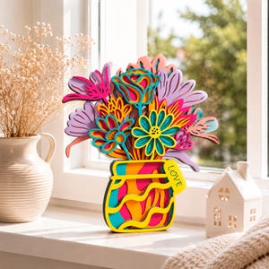 May include: A multi-layered wooden floral arrangement featuring a vibrant bouquet of stylized flowers in shades of pink, purple, yellow, and teal. The flowers are set in a matching layered vase with the word LOVE printed on a small yellow tag.