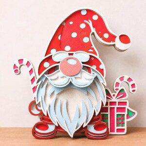 May include: A layered wooden gnome decoration featuring a red hat with white polka dots, a light blue beard, and a round nose. The gnome holds a red and white striped candy cane on the left and stands next to a red and white gift box on the right.