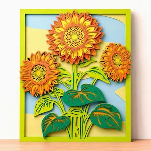 Puede incluir: Una pieza de arte mural multicapa que presenta tres girasoles con pétalos naranjas y amarillos, hojas y tallos verdes. La obra está encerrada en un marco rectangular de color verde lima brillante, sobre un fondo azul claro y amarillo pálido.
