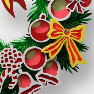 May include: A decorative wreath featuring layered cutouts of green pine needles, red and white ornaments, red bells, and a prominent yellow and red bow. The design incorporates stylized holiday elements with a multi-dimensional appearance.