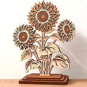 May include: A laser-cut wooden sunflower sculpture featuring three layered flowers with detailed petals and leaves, mounted on a tiered rectangular base. The design includes text labels for 1/8 in, 3 mm, 4 mm, and Digital Download.