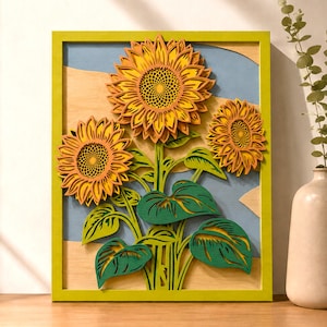 Op de afbeelding: Een meerlaags houten wandkunstwerk met drie zonnebloemen met gele en oranje bloemblaadjes, donkere harten en groene bladeren. Het kunstwerk is geplaatst tegen een lichtblauwe en natuurlijke houten achtergrond, gevat in een heldergroene lijst.