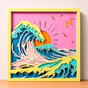 May include: A square wall art piece featuring a layered paper cut-out design of ocean waves in shades of blue, teal, and yellow against a pink sky. A bright orange sun sits behind the waves, and several orange birds are flying in the sky.