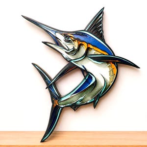 Könnte beinhalten: Ein mehrschichtiges Wandkunstobjekt, das einen springenden Marlin darstellt. Das Design besticht durch Akzente in Dunkelblau, Hellblau, Weiß und Orange mit schwarzen Konturen, was durch die sichtbaren Materialschichten einen dreidimensionalen Effekt erzeugt.