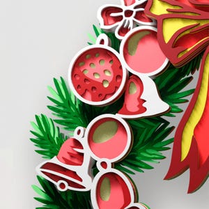 May include: A layered paper craft Christmas wreath featuring green pine needles, red and white ornaments, bells, and a large red and yellow bow. The design uses stacked cutouts to create depth, with metallic red and gold accents on the holiday decorations.
