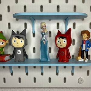 Puede incluir: Una exhibici&oacute;n de figuras en estantes azul claro fijados a una pared perforada blanca. Las figuras incluyen un elfo verde, una princesa con un vestido azul y dos figuras con trajes rojos y grises. Un libro con la palabra "CLASSICS" es visible.