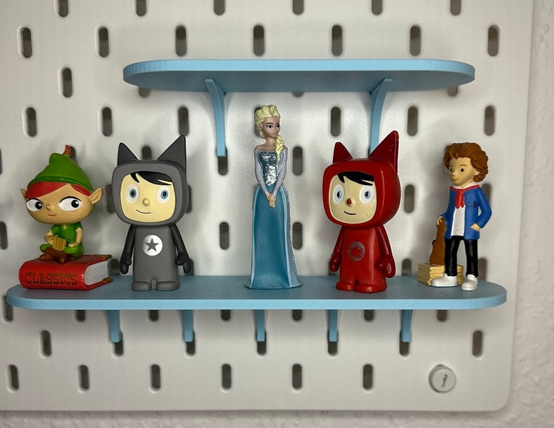 Puede incluir: Una exhibici&oacute;n de figuras en estantes azul claro fijados a una pared perforada blanca. Las figuras incluyen un elfo verde, una princesa con un vestido azul y dos figuras con trajes rojos y grises. Un libro con la palabra "CLASSICS" es visible.