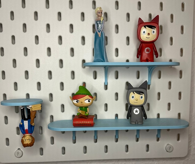 Puede incluir: Un tablero perforado blanco con estantes azules que exhiben varias figuras. Las figuras incluyen una princesa con un vestido azul, una figura roja con una estrella, una figura gris con una estrella y un elfo verde. El tablero perforado tiene agujeros espaciados.
