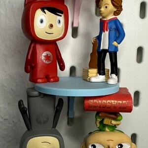 Puede incluir: Una exhibici&oacute;n caprichosa con varias figuras de dibujos animados. Una figura roja con orejas de gato est&aacute; de pie sobre una mesa azul. Otra figura con una chaqueta azul sostiene una guitarra. Otras figuras est&aacute;n boca abajo. Una mesa rosa est&aacute; en la parte superior.