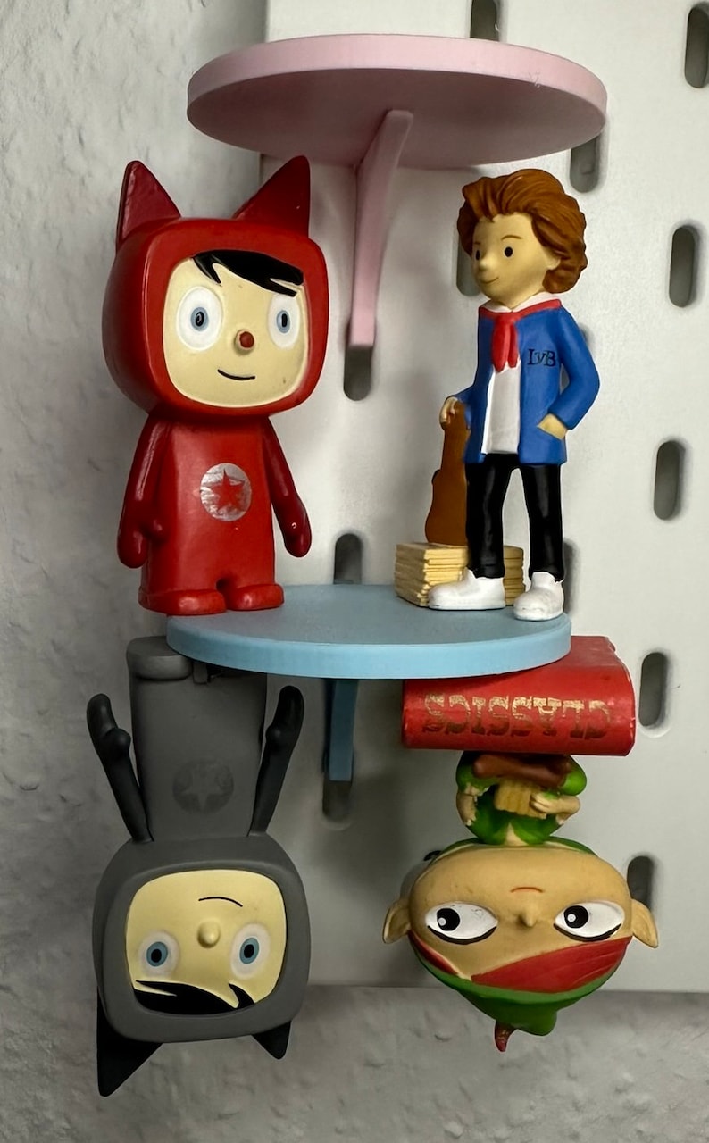 Puede incluir: Una exhibici&oacute;n caprichosa con varias figuras de dibujos animados. Una figura roja con orejas de gato est&aacute; de pie sobre una mesa azul. Otra figura con una chaqueta azul sostiene una guitarra. Otras figuras est&aacute;n boca abajo. Una mesa rosa est&aacute; en la parte superior.