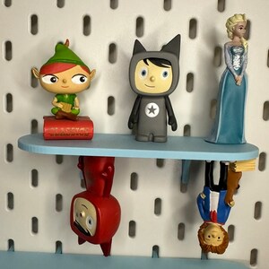 Puede incluir: Una estanter&iacute;a azul claro exhibe varias figuras: un elfo verde, una figura gris con una estrella y una princesa azul. Debajo, dos figuras m&aacute;s est&aacute;n montadas al rev&eacute;s. La estanter&iacute;a est&aacute; montada en una pared perforada blanca.
