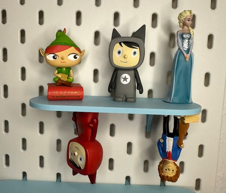 Puede incluir: Una estanter&iacute;a azul claro exhibe varias figuras: un elfo verde, una figura gris con una estrella y una princesa azul. Debajo, dos figuras m&aacute;s est&aacute;n montadas al rev&eacute;s. La estanter&iacute;a est&aacute; montada en una pared perforada blanca.