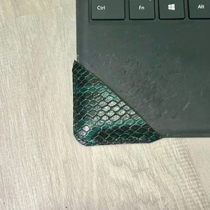 Op de afbeelding: Een groene en zwarte hoekbeschermer van leer met slangenpatroon is op de hoek van een zwart laptoptoetsenbord geplaatst. De beschermer heeft een gestructureerd oppervlak en een afgeronde rand. Het toetsenbord heeft de Ctrl-, Fn-, Windows- en Alt-toetsen zichtbaar.