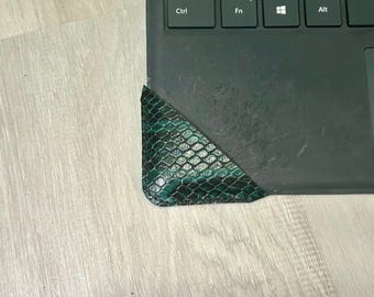 May include: A green and black snakeskin-patterned leather corner protector is placed on the corner of a black laptop keyboard. The protector has a textured surface and a rounded edge. The keyboard has the Ctrl, Fn, Windows, and Alt keys visible.