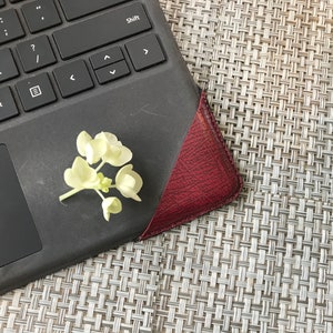 May include: A black laptop keyboard with a burgundy leather corner and a small cluster of white flowers. The keyboard is on a textured, light gray surface.