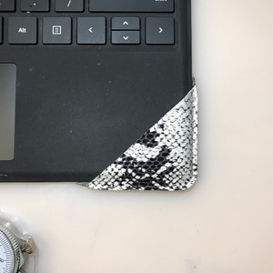 Op de afbeelding: Zwart laptoptoetsenbord met een hoekdetail met een slangenpatroon in zwart en wit. Een zilveren horloge is zichtbaar in de linker benedenhoek. Het toetsenbord ligt op een wit oppervlak.