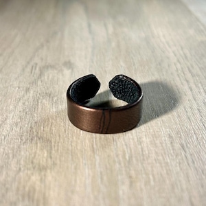 Op de afbeelding: Een bruine leren ring met een gladde, metallic afwerking aan de buitenkant en een zichtbare donkere, getextureerde voering aan de binnenkant. De band heeft een open ontwerp met twee uiteinden die elkaar niet raken, wat een opening vormt.