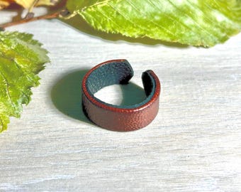 May include: A wide, open-ended ring crafted from textured leather. The exterior surface of the band is a rich, deep brown colour, while the interior lining features a contrasting dark teal or forest green hue. The band has a simple, circular shape.