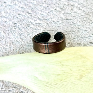 May include: A brown leather ring with a dark green interior and two thin, vertical green stripes. The ring is open at the top and sits on a light yellow surface against a textured, white background.