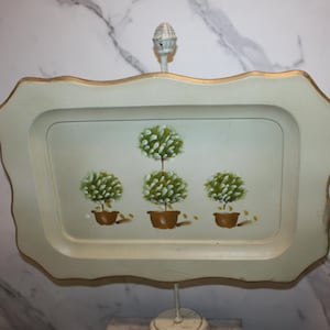 May include: A decorative tray with a scalloped edge and gold trim, supported by a white stand. The tray features a painted design of four green topiary trees in brown pots, adding a touch of elegance to any space.