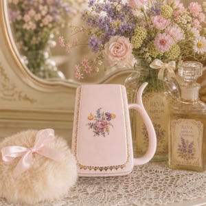 May include: A light pink ceramic mug with a floral decal and gold trim, a fluffy beige powder puff with a pink satin ribbon, and two glass bottles labeled Reva Bath Oil with floral illustrations, all arranged on a white lace doily.