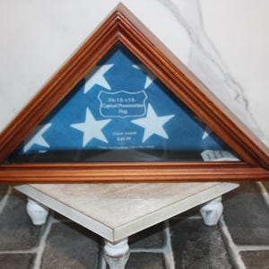 Könnte beinhalten: Dreieckiger Holz-Fahnendisplay-Kasten mit Glasfront. Der Kasten enthält eine blaue Flagge mit weißen Sternen. Der Text auf der Flagge lautet "Fits 3x5 ft. Capitol Presentation Flag". Der Kasten wird auf einem weißen Ständer präsentiert.