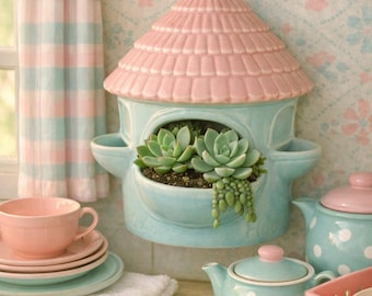 May include: A ceramic wall-mounted planter shaped like a small house with a pink roof and a light blue base. The planter holds two green succulent plants. The house-shaped planter is positioned above a kitchen sink with various dishes.
