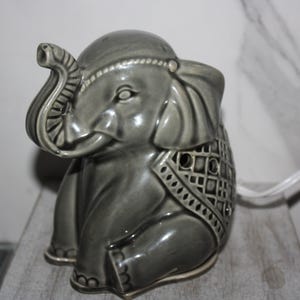 May include: A gray ceramic elephant figurine with detailed trunk and ear features. The elephant has a decorative blanket design on its back and is sitting down. The figurine has a glossy finish and is on a wooden surface.