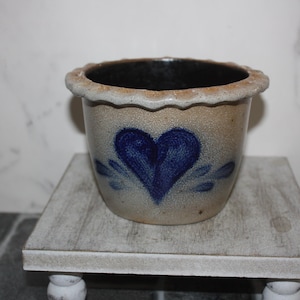 May include: A small, beige ceramic pot with a blue heart design. The pot has a scalloped rim and is displayed on a small, white, wooden stand. The interior of the pot is dark brown.