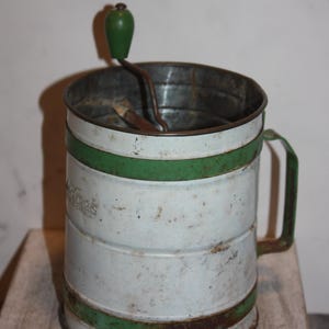 Op de afbeelding: Vintage Madrock zeef in wit en groen. De metalen zeef heeft een cilindrische vorm met groene banden en een handvat. De zeef wordt waarschijnlijk gebruikt om te bakken of te koken.