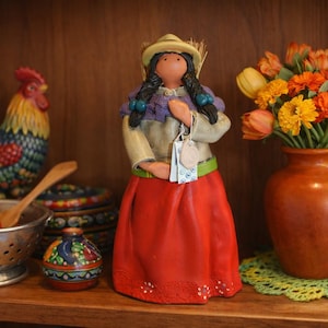 May include: A ceramic figurine of a person wearing a tan hat, a purple shawl, a light-colored top, and a long red skirt with white floral details. The figure has long black braided hair and holds a small white tag with blue patterns in their hands.