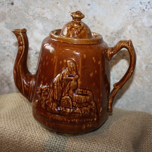 May include: A brown ceramic teapot with a lid and a handle. The teapot has a raised design of a woman drawing water from a well.