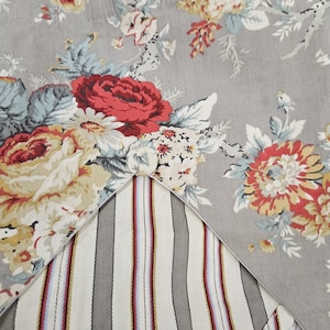 Puede incluir: Primer plano de una tela floral con fondo gris. La tela presenta grandes rosas rojas, amarillas y blancas con hojas verdes y azules. Se ve una sección de tela a rayas con rayas grises, rojas y azules.