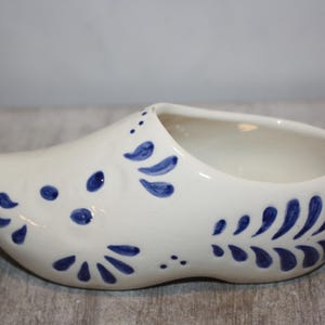 May include: White ceramic clog-shaped planter with blue floral designs. The planter has a rounded shape with an open top for planting. Decorative blue accents adorn the sides and front of the clog.