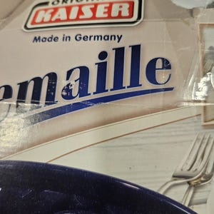 May include: Close-up of a box with the text "ORIGINAL KAISER Made in Germany." The box is beige with blue text and a red and white logo. A dark blue object is visible at the bottom of the image, along with a silver fork.