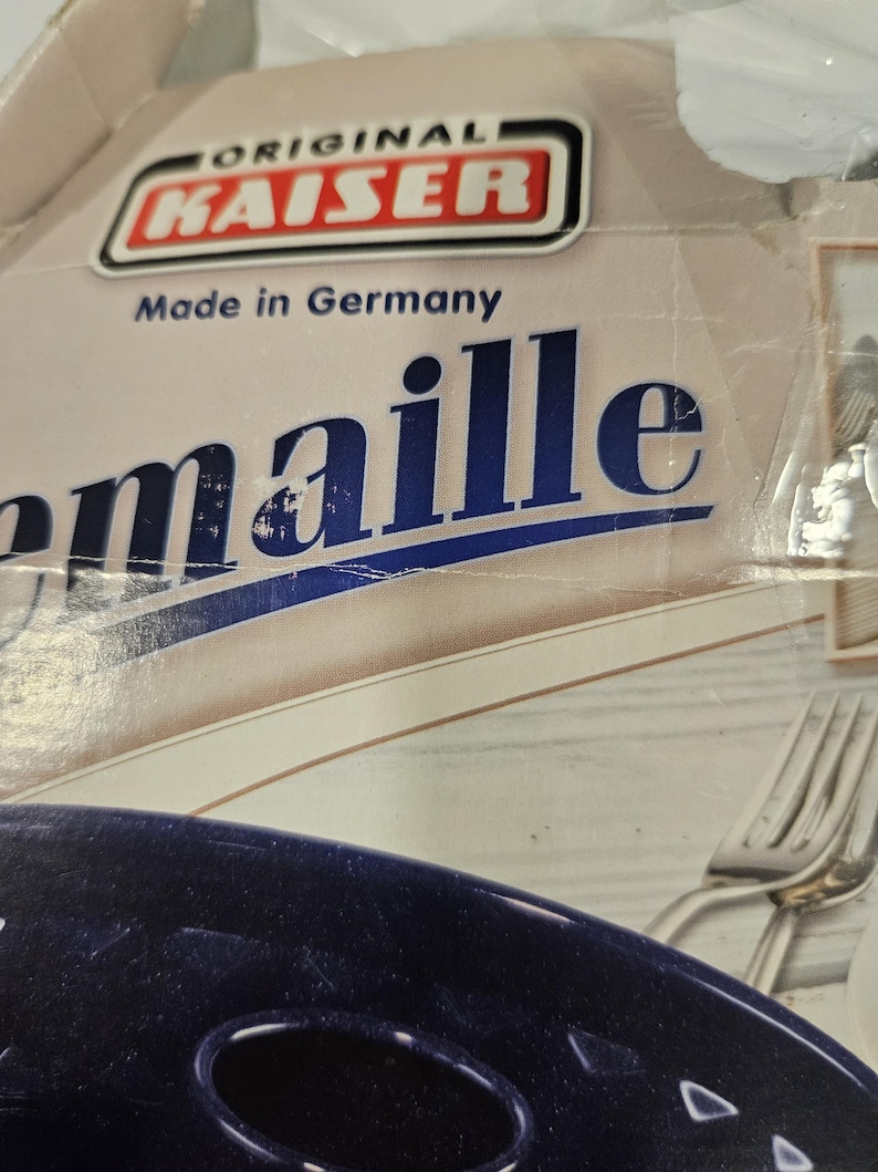 May include: Close-up of a box with the text "ORIGINAL KAISER Made in Germany." The box is beige with blue text and a red and white logo. A dark blue object is visible at the bottom of the image, along with a silver fork.