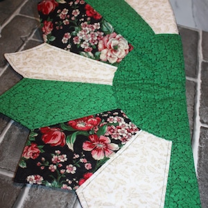 May include: A quilted table runner with a green leaf pattern, accented with floral fabric in black and cream. The floral fabric features red and pink roses and small white flowers. The runner is made of multiple fabric pieces.